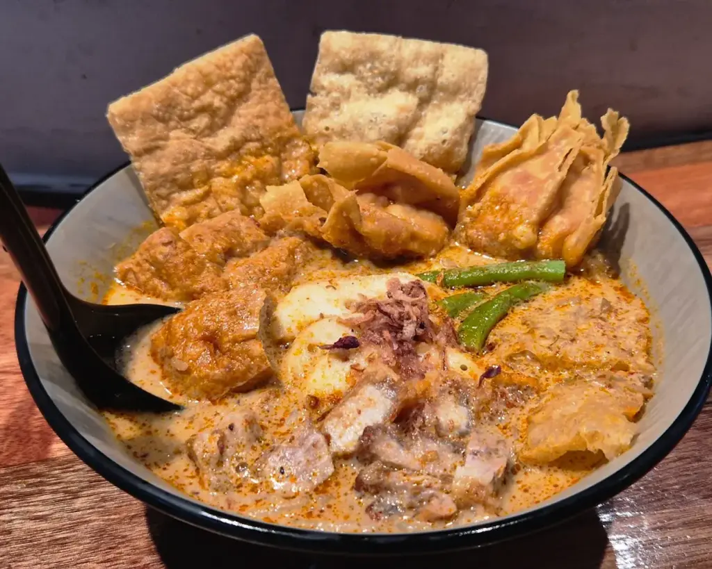 A hearty bowl of curry mee with rich, coconut-based broth, noodles, tofu, seafood, and herbs, offering a comforting and aromatic local favourite.
