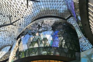 A modern building, Ion Orchard, Singapore, featuring a glass roof and a large reflective mirror on its facade.