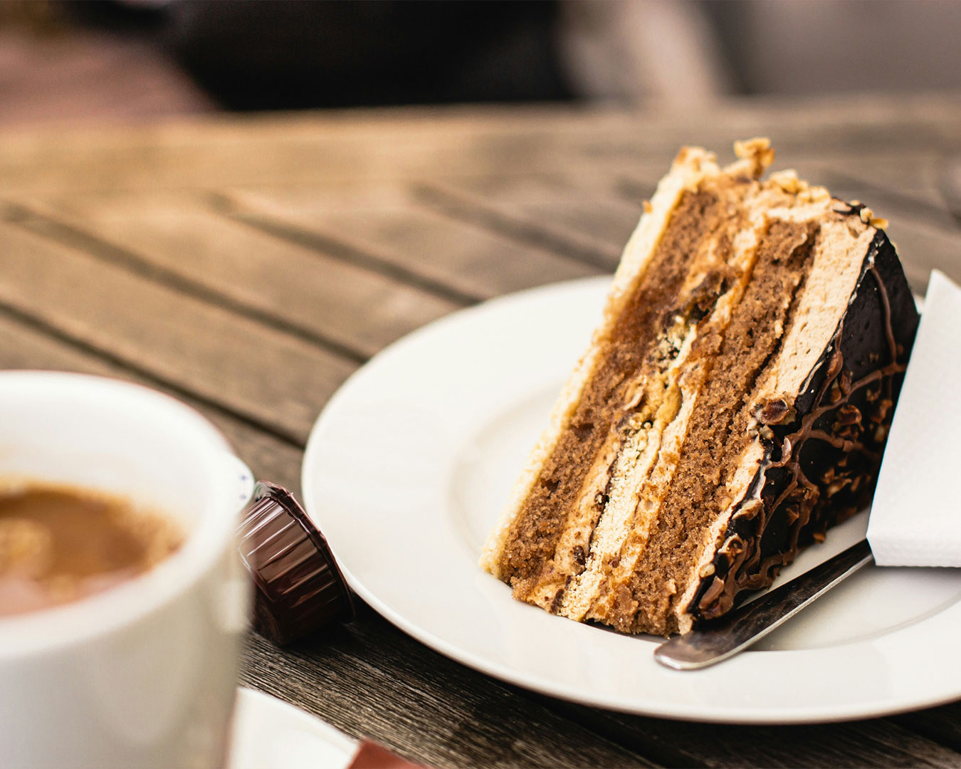 A rich slice of chocolate cake served on a plate beside a cup of hot coffee, with smooth chocolate frosting and visible layers, creating a cosy café-style dessert setting