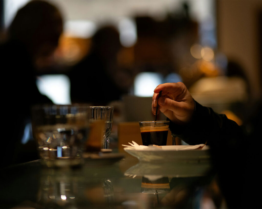 Close-up of a hand holding a glass of iced drink inside a restaurant, with softly blurred tables and warm ambient lighting creating a relaxed dining atmosphere