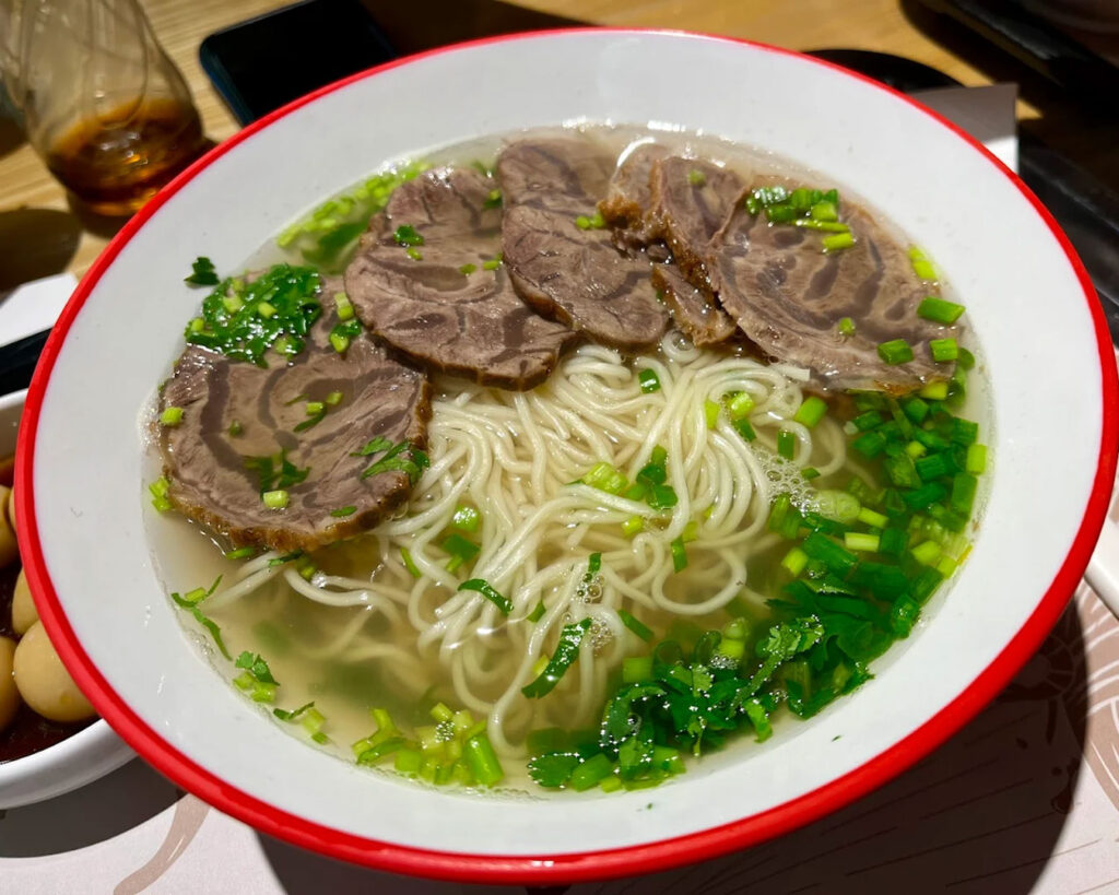 A warm bowl of noodle soup with clear broth, topped with vegetables and slices of meat, giving a comforting and hearty feel.