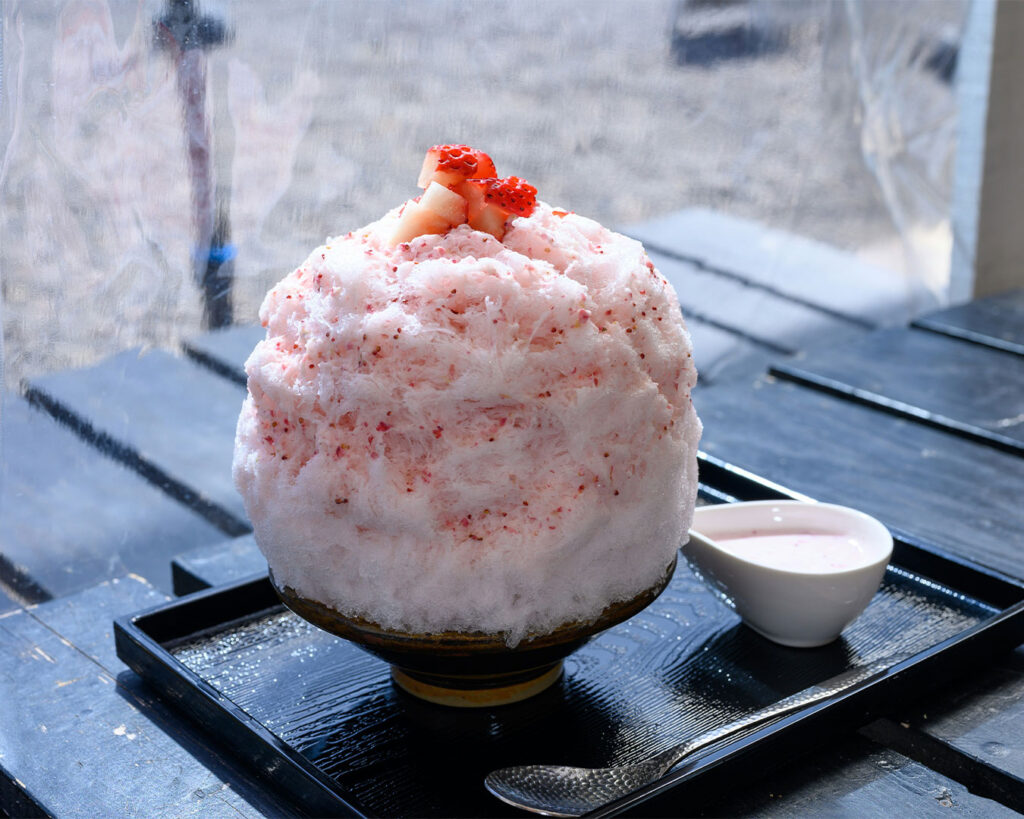 A bowl of colourful shaved ice dessert topped with syrup and fruit, refreshing and bright against a simple background.