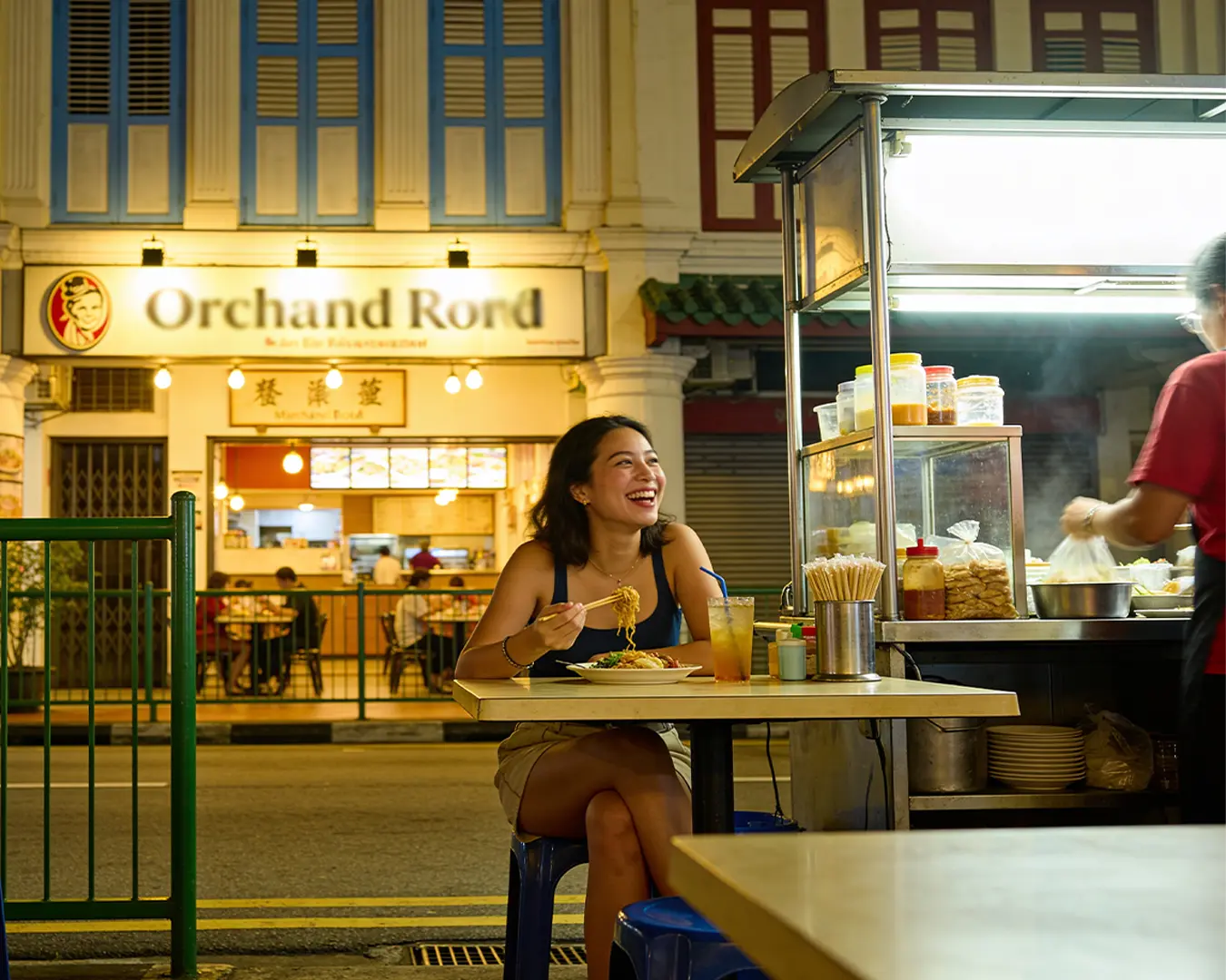 A woman sitting at an outdoor dining table along Orchard Road at night, enjoying her meal while the city lights, and illuminated storefronts create a lively urban dining atmosphere.