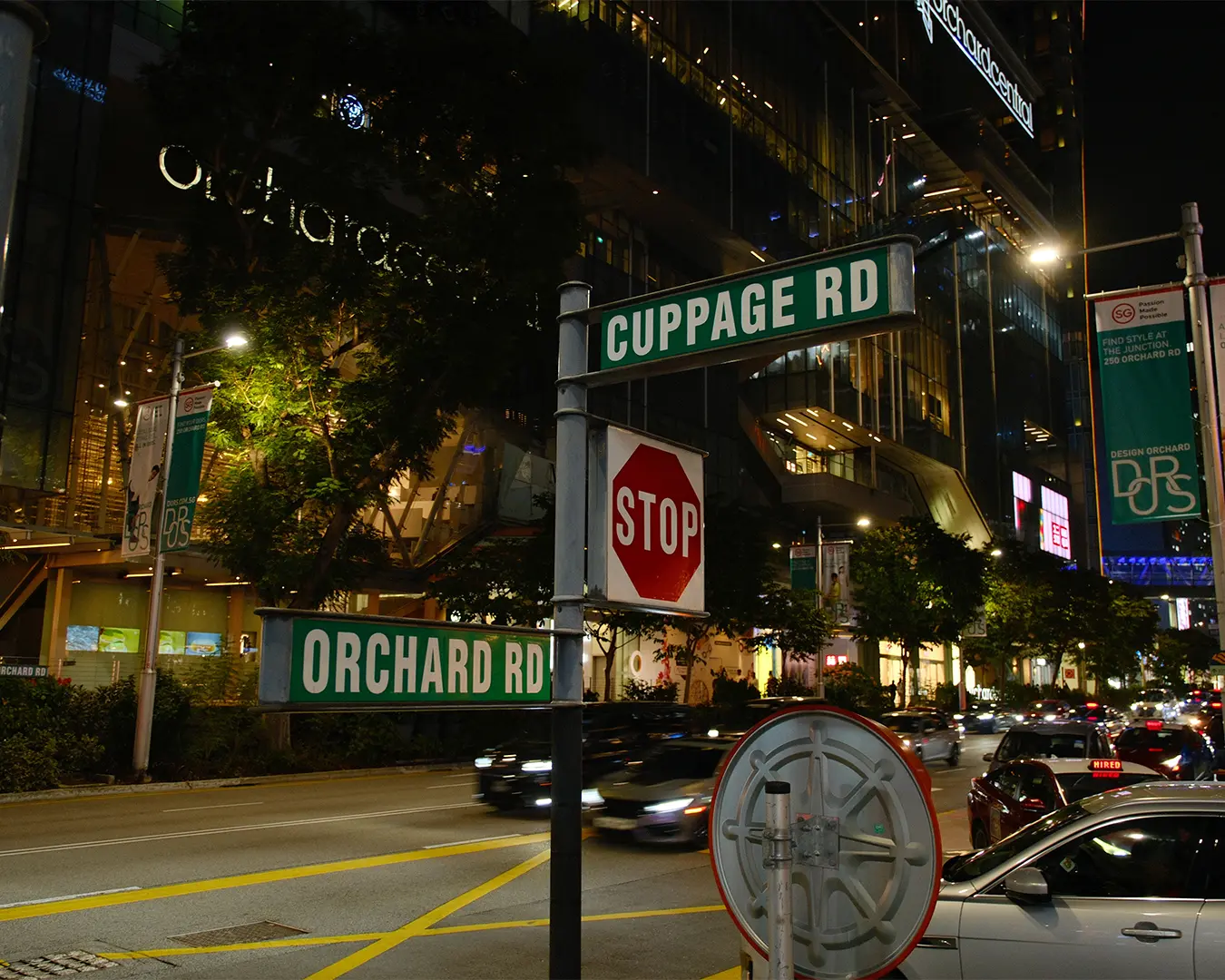 The illuminated Orchard Road street sign glowing at night, surrounded by city lights, moving traffic, and modern shopping malls that highlight the lively atmosphere of Singapore’s famous shopping boulevard.