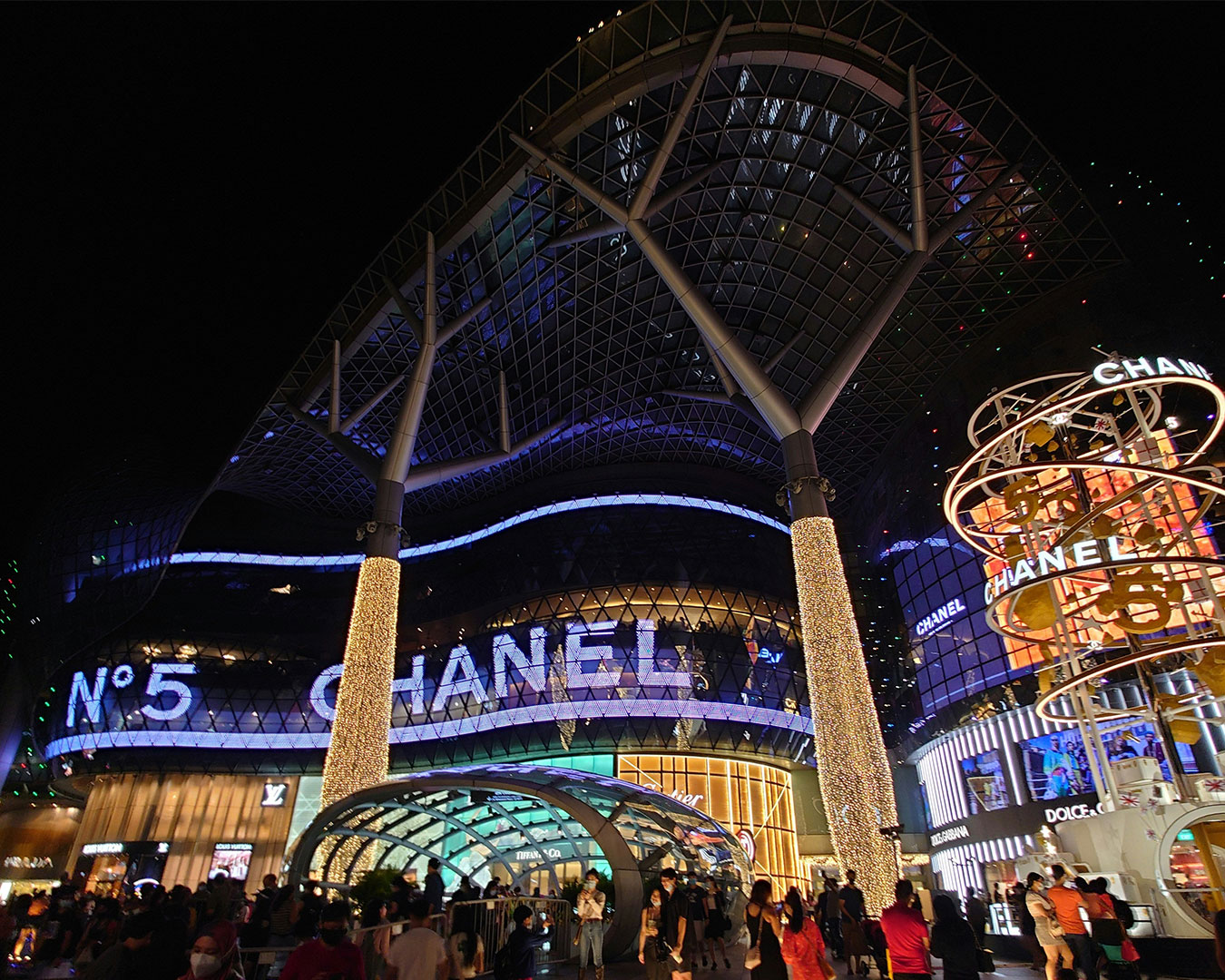 A large modern shopping mall along Orchard Road, featuring glass façades, bright storefronts, and luxury brand signage, with pedestrians visible in the foreground.