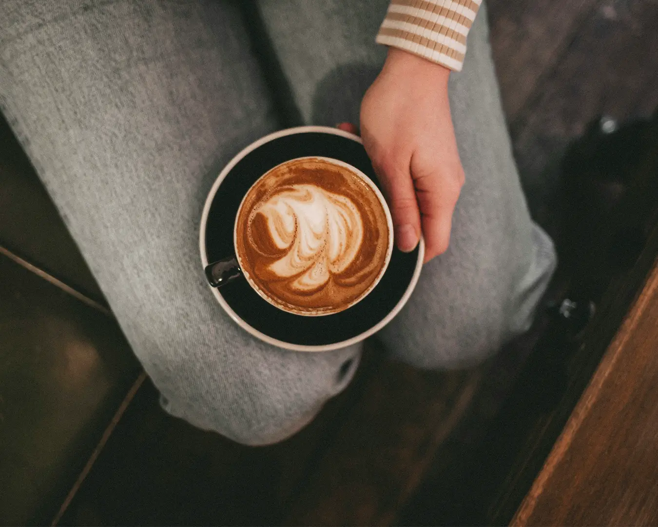 A person gently holding a warm cup of coffee while it rests on their lap, creating a calm and cosy moment. The soft steam rising from the cup adds to the relaxed atmosphere, suggesting a quiet break or a peaceful morning ritual.