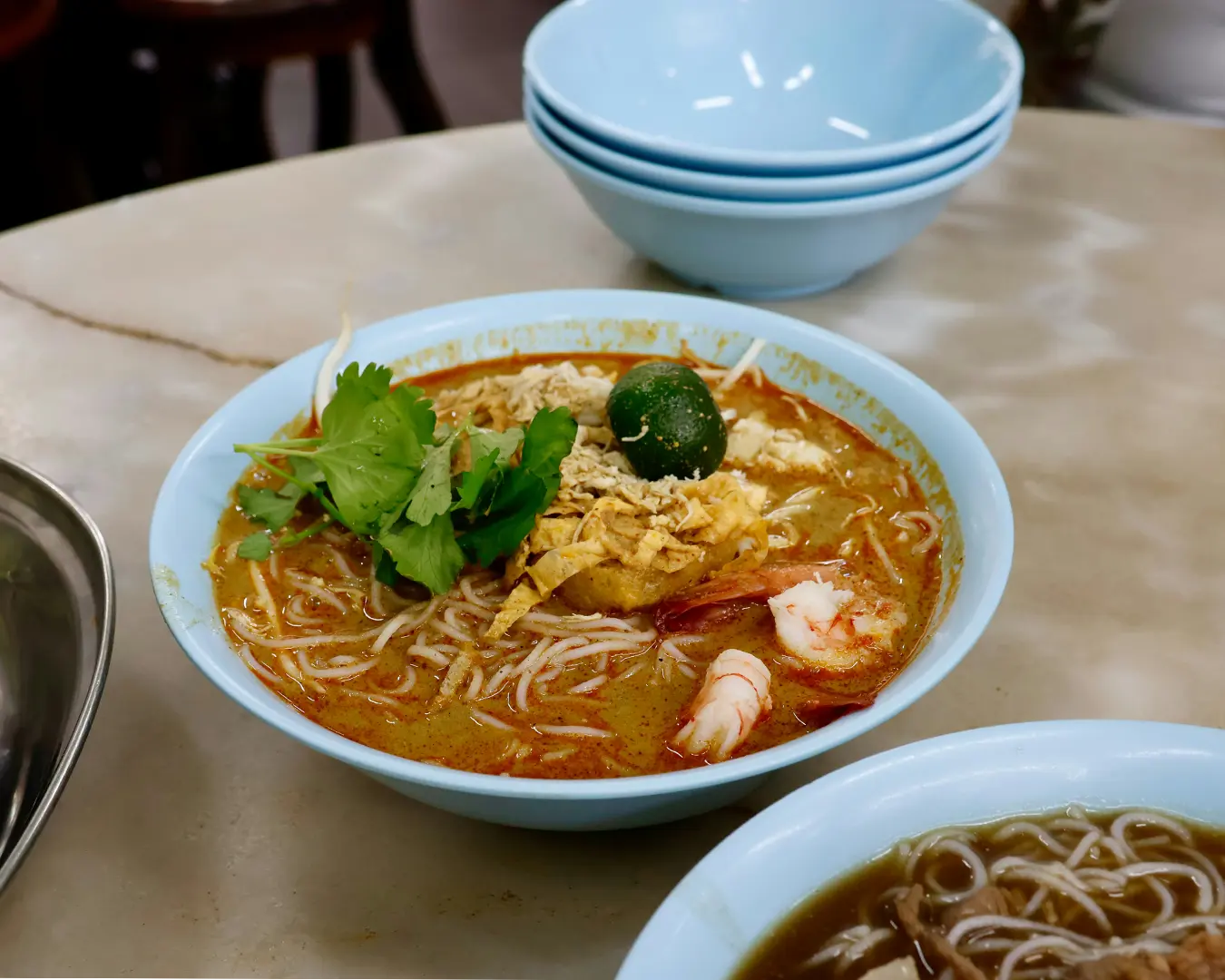 A bowl of rich curry laksa with noodles, prawns, and tofu puffs served on a dining table, with the spicy coconut broth visible.