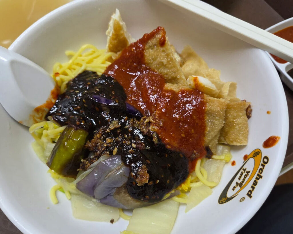 A bowl of yong tau fu filled with assorted tofu, vegetables, and fish paste items in clear broth, served hot with visible steam and garnished simply for a comforting, homestyle look.