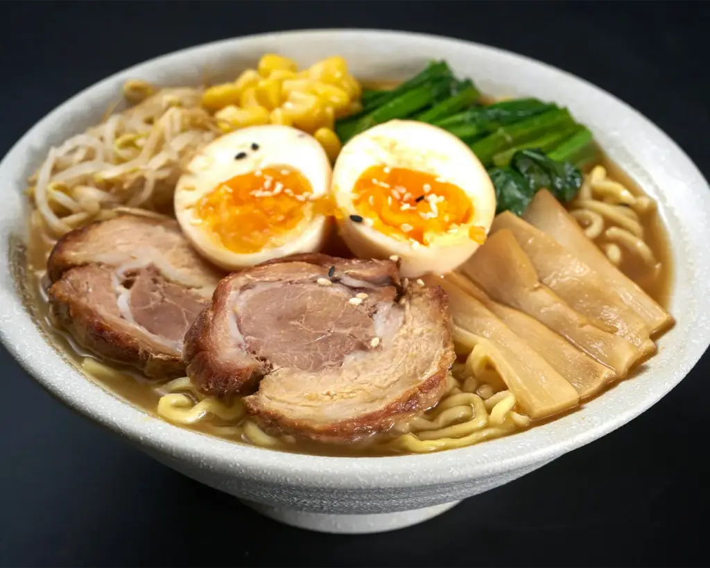 Close-up of a steaming bowl of ramen with rich broth, noodles, sliced meat, soft-boiled egg, and garnishes, presented in a restaurant setting with warm and inviting lighting.