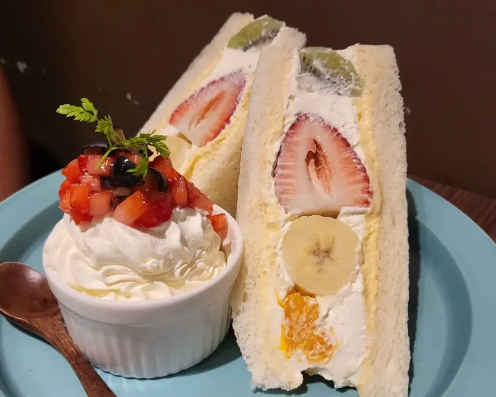 A sliced bread dessert filled generously with fresh cream and colourful fruits including strawberry, mango, kiwi, and banana. The soft bread and bright fruit layers create a sweet and refreshing treat that looks both light and indulgent.