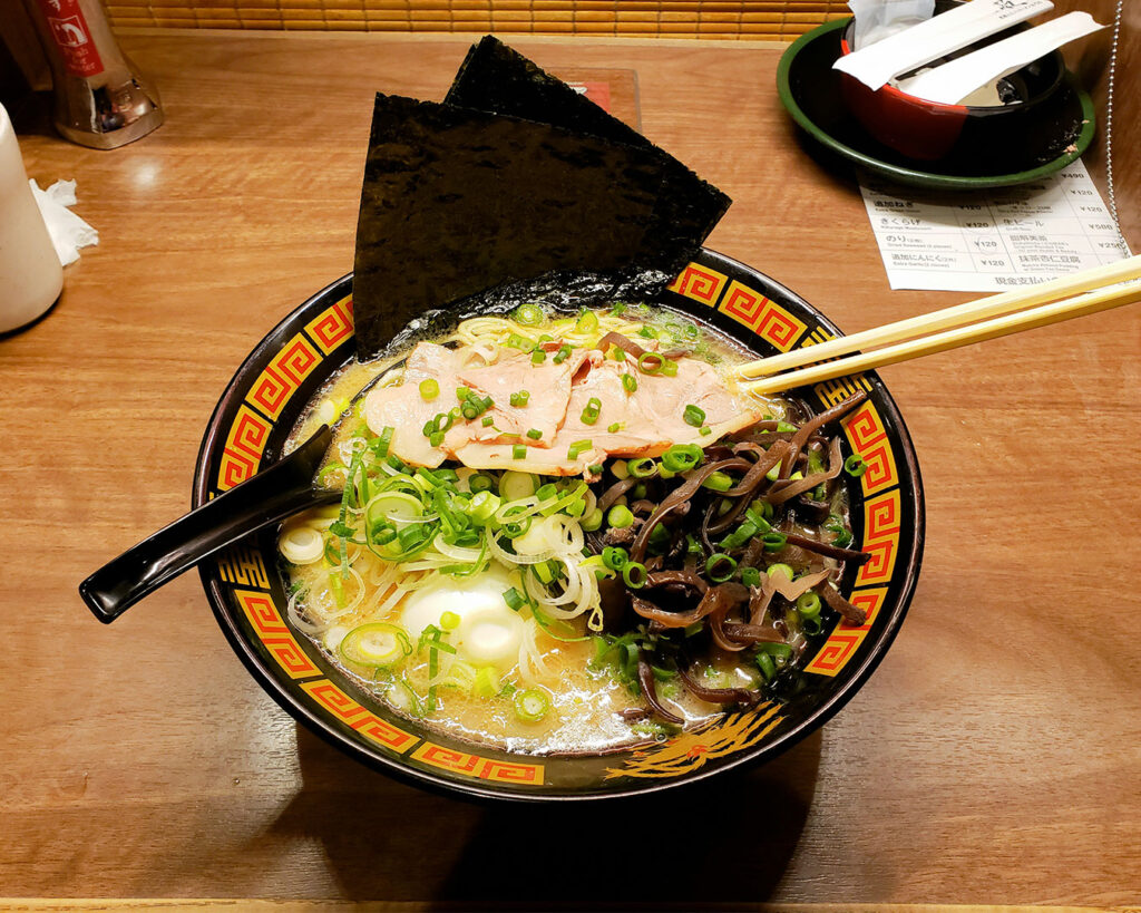 A bowl of ramen placed on a wooden table, topped with sliced meat, soft-boiled egg, and spring onions, with rich broth and noodles visible under warm restaurant lighting.