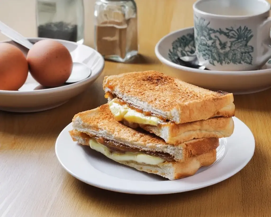 A traditional Kaya toast set with toasted bread spread with kaya coconut jam and butter, served alongside a cup of Kopi in a classic local café setting.