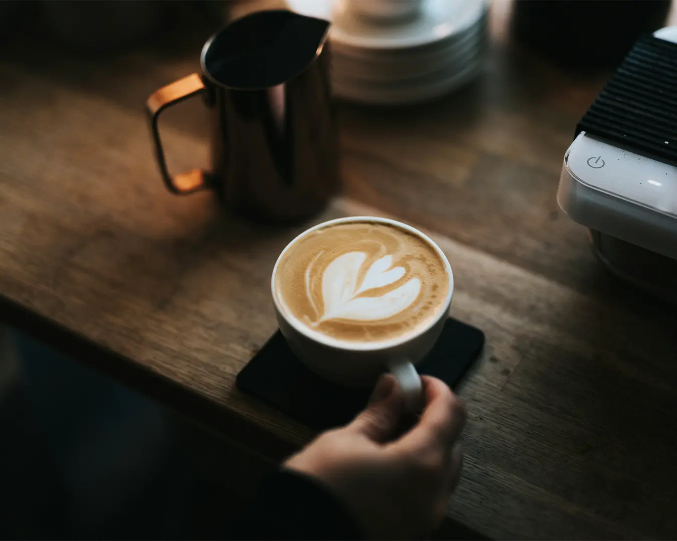 A hand holds a white cup of latte with intricate heart-shaped latte art on a wooden table. A metal pitcher and espresso machine are in the background, exuding a cozy café vibe.