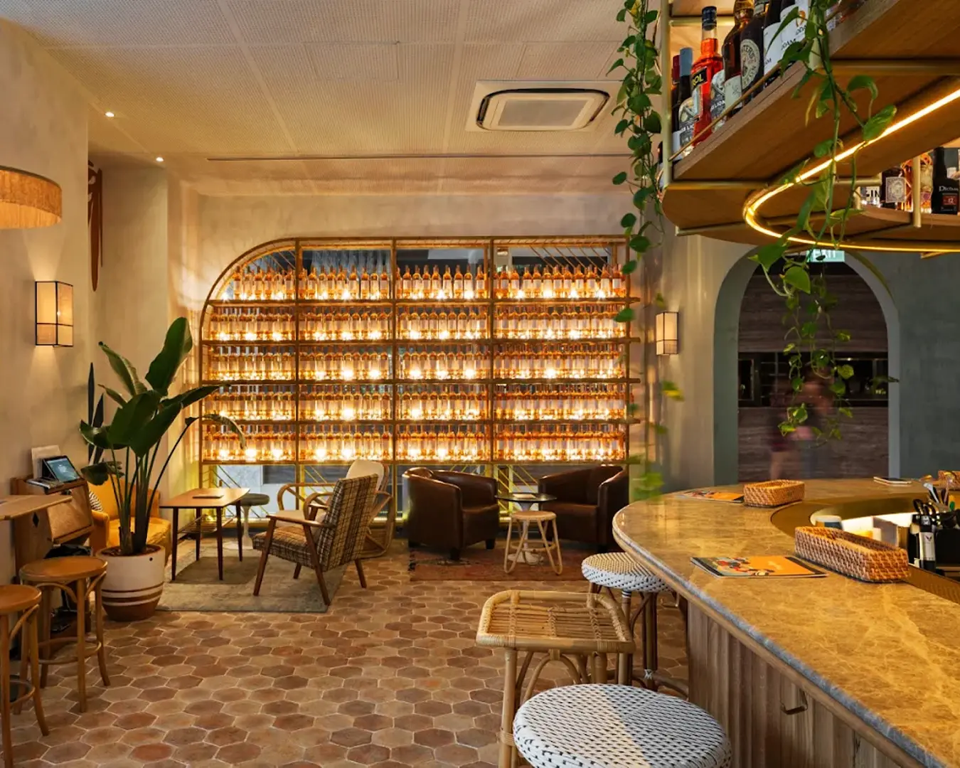 Chic bar interior with warm lighting and cozy atmosphere. Backlit shelves display numerous bottles, surrounded by wicker and leather seating, plants, and a marble counter.