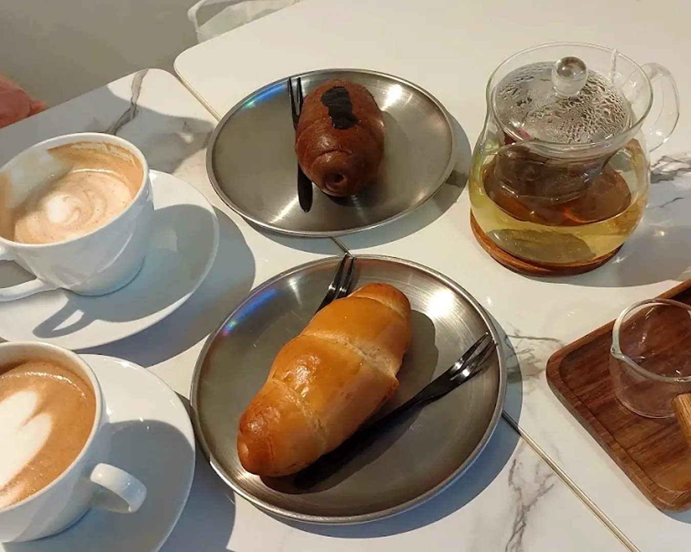 A cafe table with two cappuccinos in white cups, a croissant, a chocolate pastry on metal plates, and a clear teapot with tea, creating a cozy atmosphere.