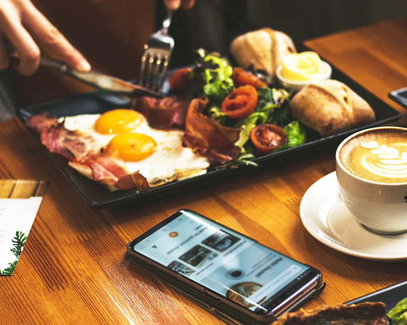 A close-up of a breakfast plate with sunny-side-up eggs, bacon, salad, and bread. A latte and a smartphone rest on a wooden table, creating a cozy, inviting scene.