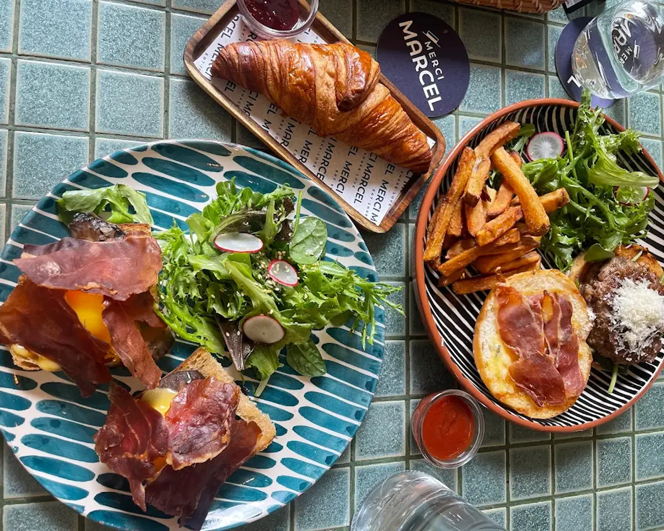 A vibrant brunch spread on a tiled table includes a grilled sandwich with crispy prosciutto, fresh greens, a croissant with jam, and a dish of sweet potato fries.