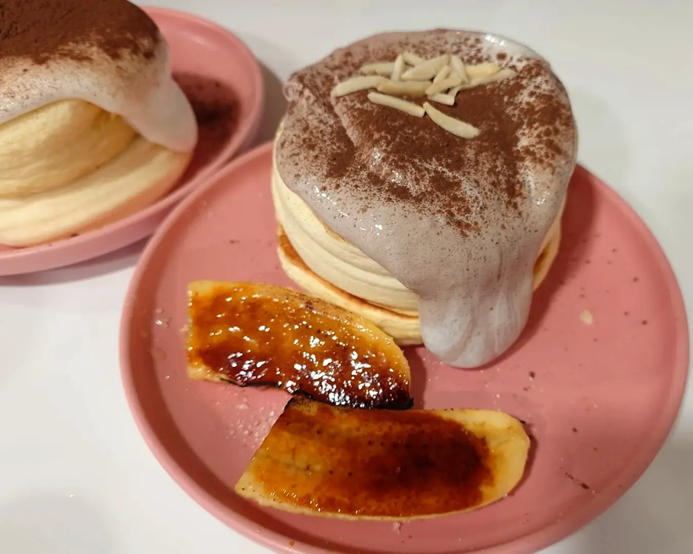 Fluffy pancakes topped with a creamy sauce and cocoa powder, garnished with almond slivers, next to caramelized banana slices on a pink plate.