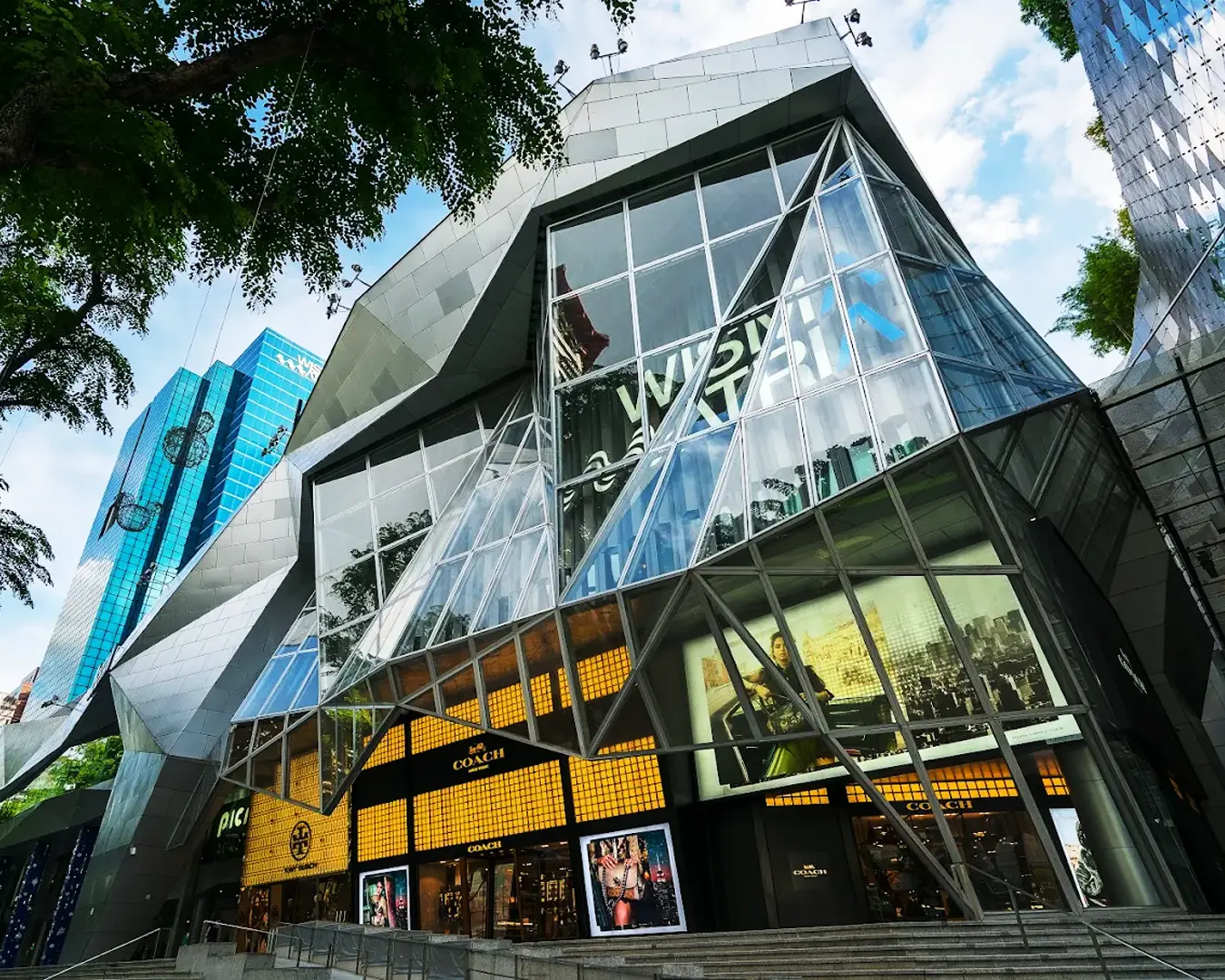 Futuristic glass and metal building with angular design, surrounded by trees. Bright shopping displays inside convey a vibrant, urban atmosphere.