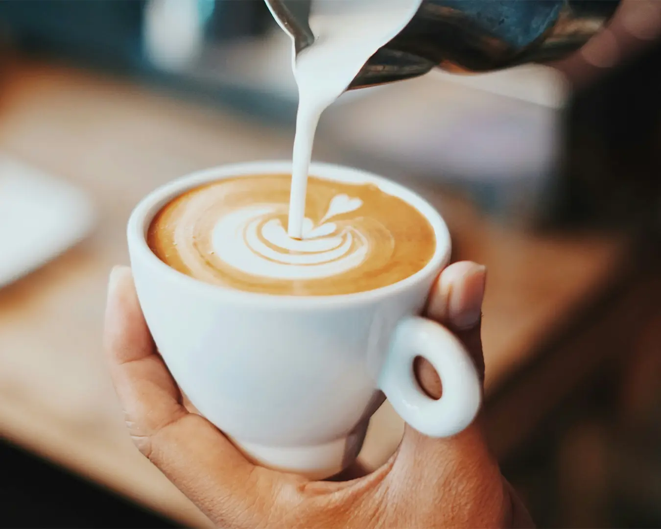 A hand holds a white cup of cappuccino as milk is poured, creating intricate latte art. The background is softly blurred, conveying a cozy café ambiance.
