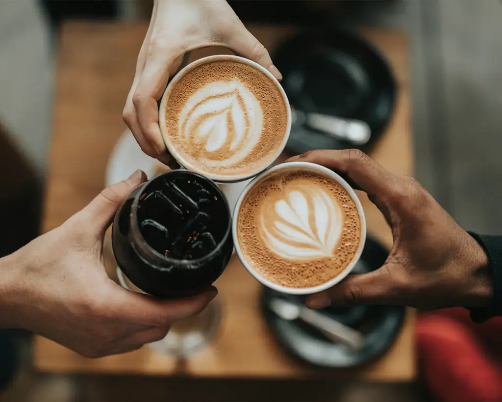 Three hands clink together, holding two cups of latte with heart-shaped foam art and a glass of iced coffee, over a wooden table. Cozy and inviting mood.